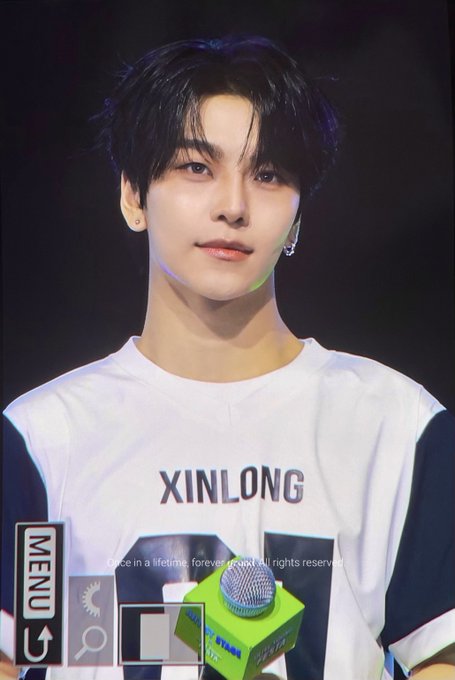 A young man with dark hair wearing a white "XINLONG" jersey holds a microphone, matching the post's tag #XINLONG (鑫隆/씬롱) referencing Alpha Drive One (ALD1/알디원). The menu icons and text overlay suggest this is a screenshot from a fan photo taken at a 2026 event.