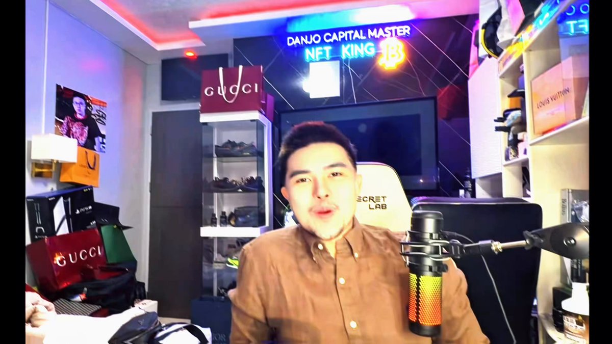 The image shows a man sitting at a microphone, likely recording a podcast or live stream. He appears to be wearing a brown shirt and has a microphone in front of him. The background features a TV screen displaying the words "Danjo Capital Master" and "NFT King 13". There are also several other items scattered around the room, including a backpack on the floor near the man's feet and various bags placed throughout the space.