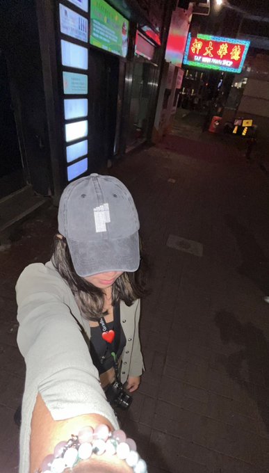 A nighttime selfie of a person in a gray cap and white cardigan, with a "I ❤️" t-shirt, standing on a city street near a red sign reading "TAY HING HOUSE LTD" and Chinese characters.