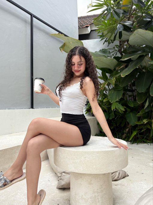 A photograph of a young woman with long curly brown hair, wearing a white tank top and black skirt, sitting on a stone bench and holding a white paper coffee cup in an outdoor area with green plants.