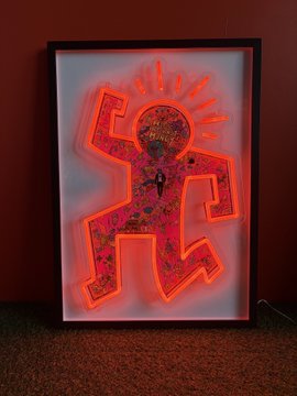 A framed neon artwork depicts Keith Haring's iconic radiating baby figure in glowing red and pink, casting light onto the floor beneath it.