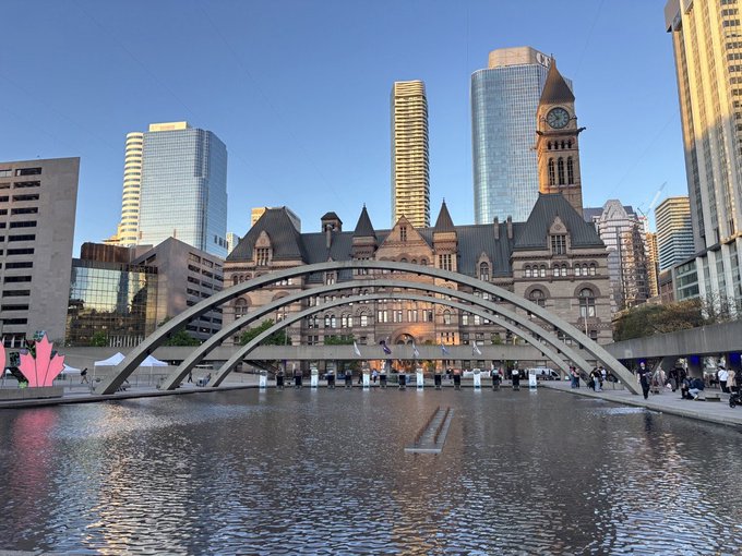The image shows a large body of water with a bridge spanning across it. The bridge has an arch design and spans the width of the river. In front of the bridge, there are buildings that contribute to the cityscape. A clock can be seen on one of these buildings. The scene is set against a clear blue sky, creating a picturesque view of the city's architecture and natural elements.