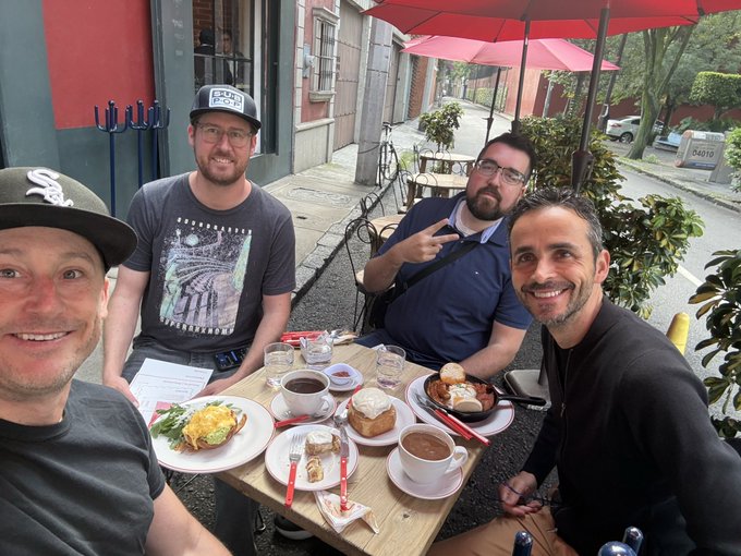 Four men sit together at an outdoor café table under a red umbrella, smiling for a photo while sharing brunch dishes including eggs, sandwiches, cake, and coffee.
