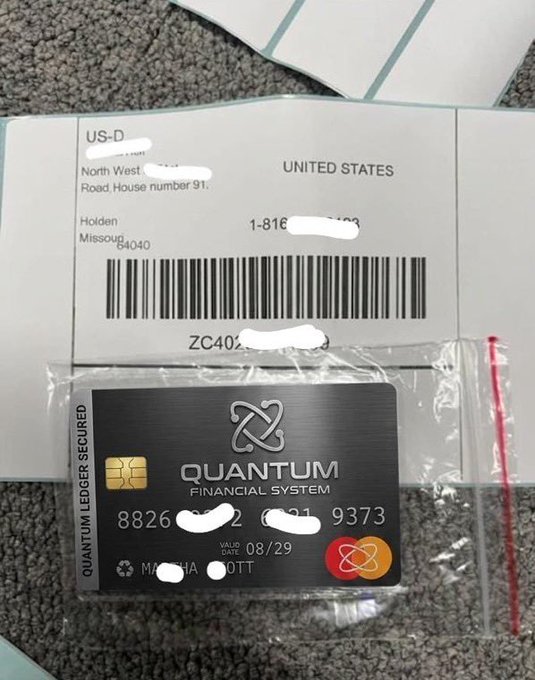Photograph of a delivery package containing a credit card from "Quantum Financial System" with a metal design, Mastercard logo, and a US shipping label with personal details redacted.