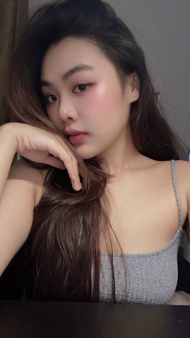A selfie-style portrait of a young woman with long dark hair wearing a gray tank top, resting her chin on her hand with makeup including pink blush and lip color.