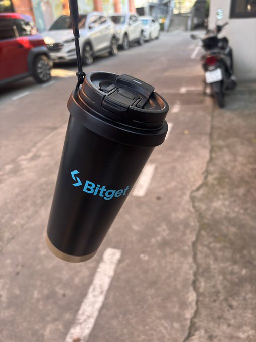A black Bitget-branded travel tumbler hanging from a strap against a blurred street background with parked cars and a motorbike.
