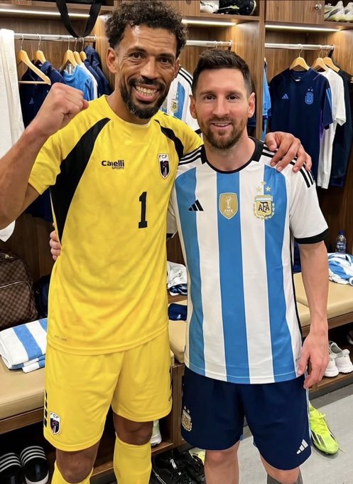 A photo of two men smiling and posing in what appears to be a sports locker room with team jerseys hanging in the background. The man on the left wears a yellow goalkeeper uniform with the number 1, while the man on the right wears an Argentina national team jersey with light blue and white stripes.