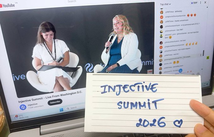 A handwritten sign reading "INJECTIVE SUMMIT 2026 ♡" is held up in front of a YouTube livestream of the event, showing two women on stage at the Washington D.C. summit (77°F) with an active chat sidebar. The image captures a fan attending the highly anticipated onchain finance gathering virtually alongside notable figures from the U.S. government and financial institutions.