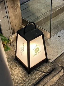 A black-framed Take Me Out Cafe lantern with a warm glow sits on stone steps outside a glass-fronted entrance.