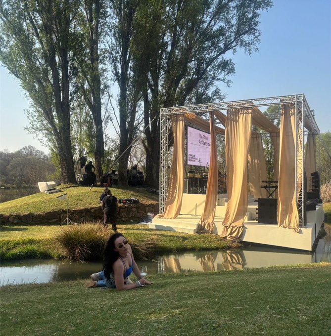A woman relaxes on the grass near a tranquil pond in front of a "Web3 Art Experience" event stage, embodying the post's irony of trying to escape Web3 by going outdoors but still encountering it.