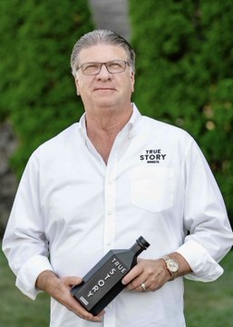 A man with gray hair and glasses, wearing a white "TRUE STORY" branded shirt and holding a black bottle labeled "25 MILLION," stands outdoors in front of greenery. The image relates to the article as it appears to feature Wes Henderson, the bourbon mogul mentioned in the headline, who is reportedly associated with "True Story" branding.