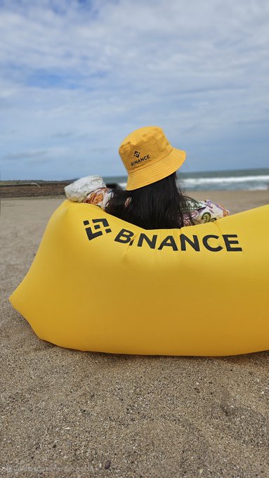 A person wearing a yellow Binance bucket hat relaxes on a matching yellow Binance-branded inflatable lounger on the sandy beach, with the ocean and cloudy sky in the background—capturing a leisurely weekend ("fds") vibe.