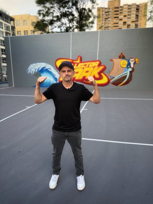 A man in a black t-shirt, jeans, and red cap flexing his biceps on an outdoor court with colorful graffiti on the wall behind him.