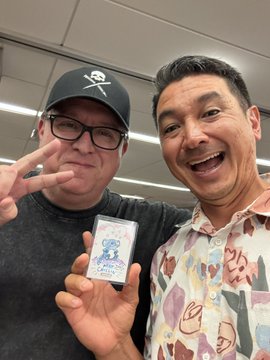 A selfie of two smiling men; the man on the left wears a black cap with a skull logo and glasses, holding a Pokémon card, while the man on the right wears a colorful patterned shirt.