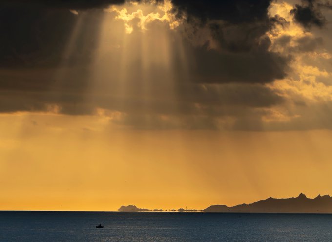A dramatic seascape at sunset with golden sunbeams piercing through dark clouds over the ocean, where a small boat floats near a distant silhouetted island.