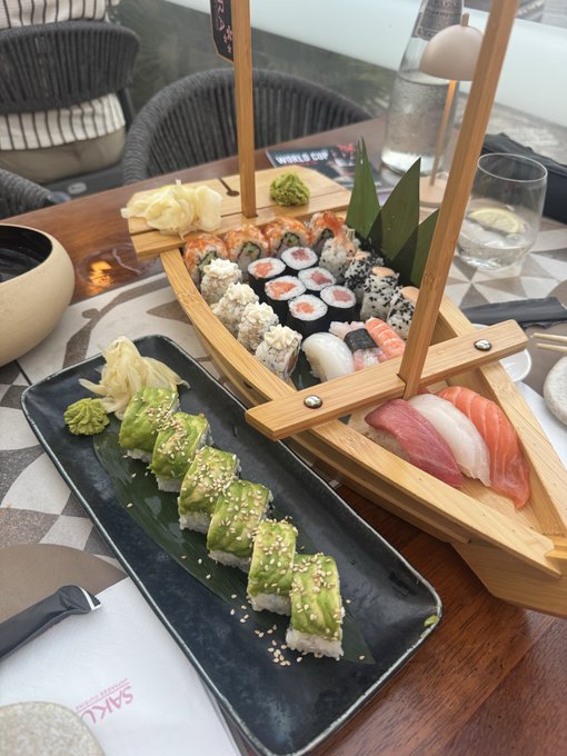 A boat-shaped wooden platter showcases an assortment of sushi rolls and sashimi (salmon, tuna, and white fish), paired with a separate plate of avocado-topped rolls sprinkled with sesame seeds. The presentation is elegant, with pickled ginger and wasabi on the side, and a "SASA" branded napkin visible, making it a tempting "smash" for any sushi lover.