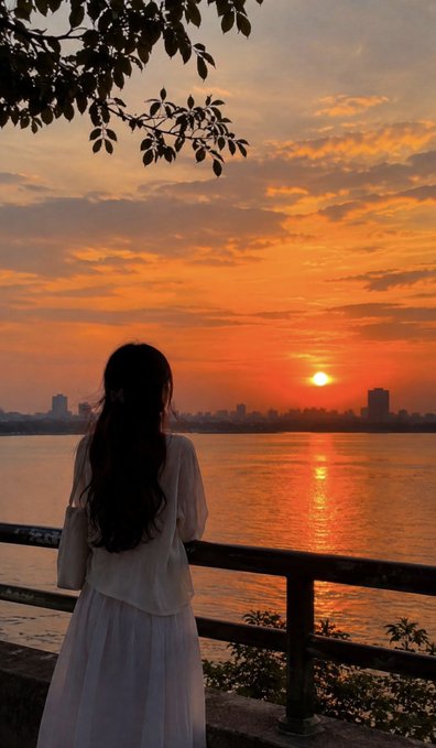 A woman in a flowing white dress stands at a railing watching a vibrant orange sunset over a river with a city skyline in the distance.