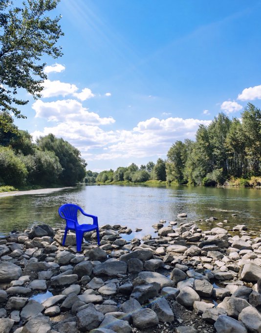 A serene landscape showing a lone blue plastic chair sitting on a rocky riverbank beside a calm river, surrounded by trees under a partly cloudy blue sky.