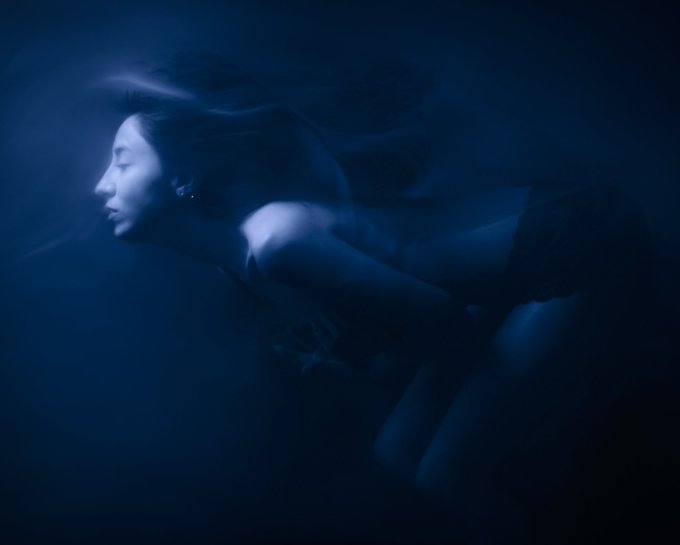 The image features a woman with long hair, wearing black shorts, captured in a moment of stillness or rest. The background appears to be dark blue, which contrasts with her hair color. The photo has a slightly blurred effect, giving it an artistic and dreamy quality. The caption "Blue Mood" suggests that the image is related to the concept of mood or atmosphere, possibly evoking feelings of tranquility or introspection.