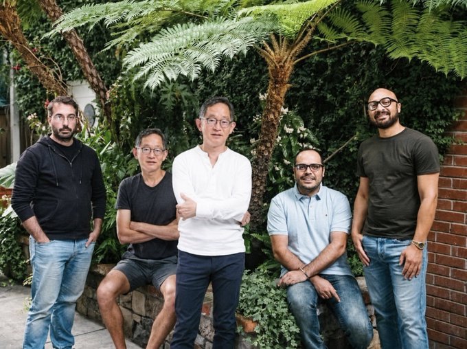A group of five men pose together outdoors in front of lush green tree ferns and a brick wall, dressed casually in t-shirts, sweaters, and jeans.