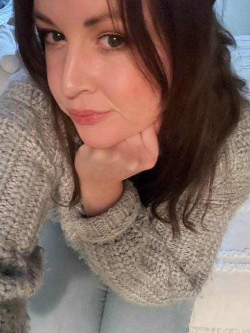 A close-up selfie of a woman with dark brown hair wearing a gray knit sweater, resting her chin on her hand with a slight smile.