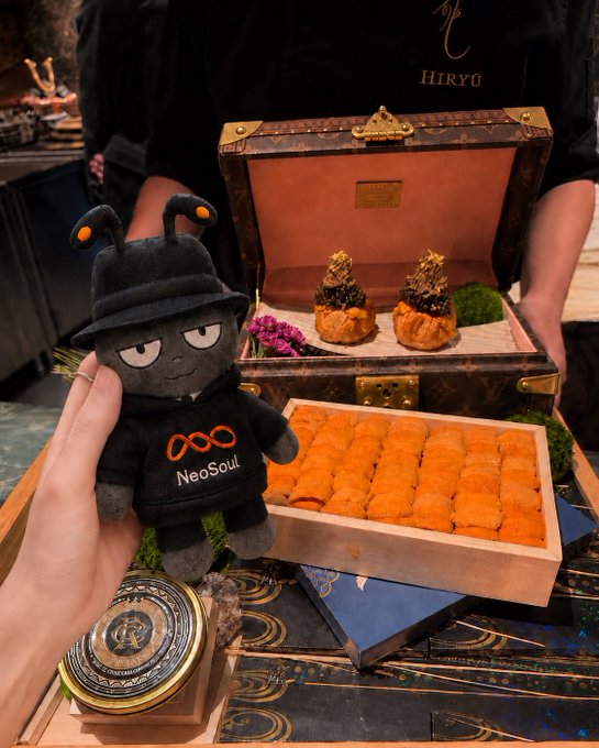 A plush 'NeoSoul' mascot wearing a black outfit stands next to an ornate HIRYU presentation box filled with golden uni (sea urchin) and luxurious sushi on a decorative tray.