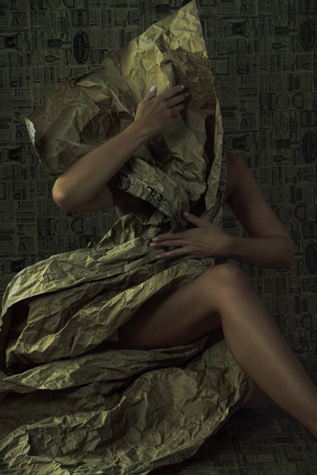 An artistic photograph of a figure wrapped in crumpled gold paper, obscuring their face with their hands against a wall covered in newspaper print.