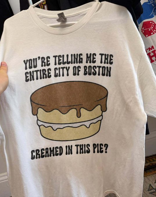 A cream-colored t-shirt is displayed on a hanger, featuring a cartoon illustration of a Boston cream pie with the humorous text: "YOU'RE TELLING ME THE ENTIRE CITY OF BOSTON CREAMED IN THIS PIE?"