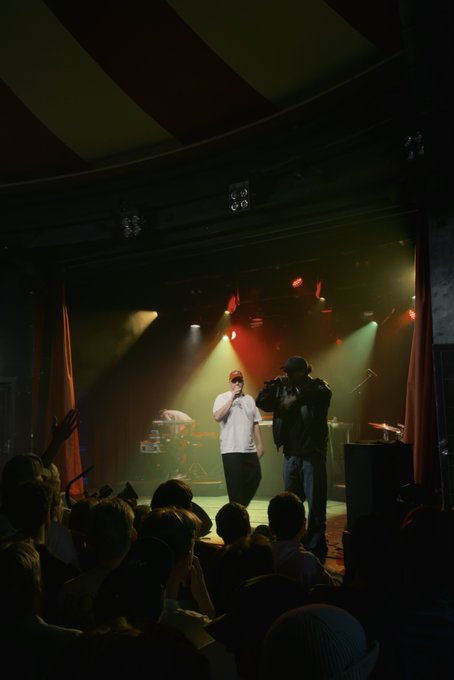 A live concert performance unfolds on a dimly lit stage bathed in dramatic red and white spotlights, where an artist in a white t-shirt and cap holds a microphone while another person films him, with an enthusiastic crowd raising their hands in the foreground—capturing the intimate underground venue atmosphere associated with the Tøyen Holding event.