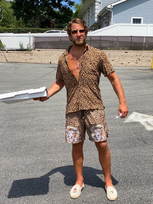 A bearded man wearing a leopard print outfit with cherub-printed shorts holds pizza boxes in a parking lot, humorously matched to the post's warning about not letting him "drop any more coins," suggesting his quirky style makes him seem unreliable or distracted.
