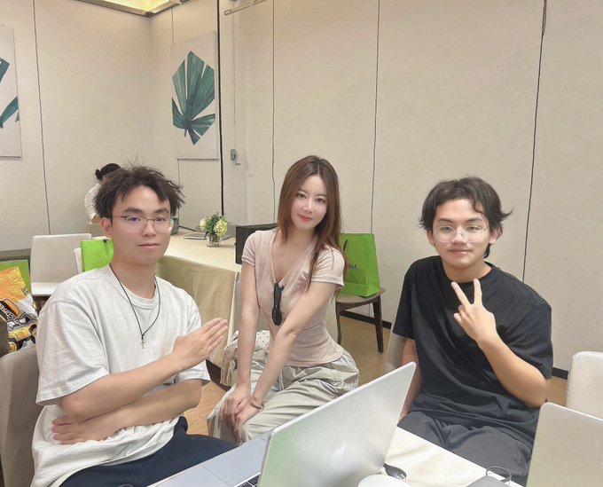 Three young people pose together indoors at what appears to be a meetup event, with two men on either side flashing peace signs while a woman sits smiling in the middle, all seated behind laptops in a casual room with decorative greenery on the wall. The image illustrates the post's mention of an in-real-life gathering where the author met up with fellow crypto/Web3 community members at the Xclub Shenzhen event.