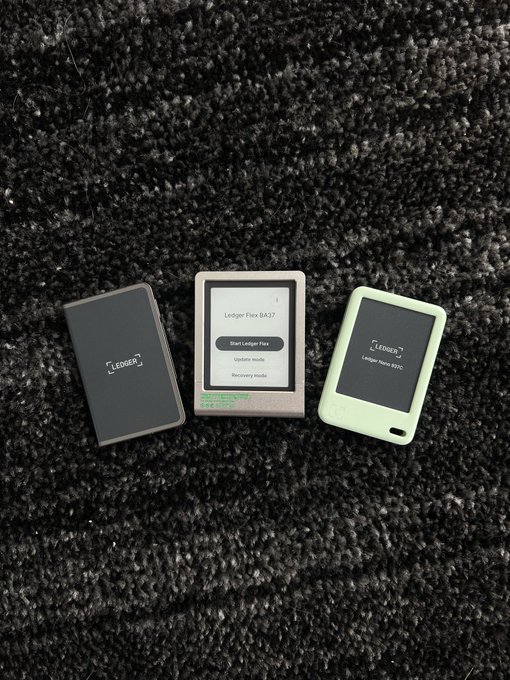 Three Ledger hardware wallets are displayed on a dark textured surface, featuring the "Ledger Flex" (BA37), "Ledger Nano X," and a model displaying its initial setup screen with "Start Ledger Flex," "Update mode," and "Recovery mode" options.