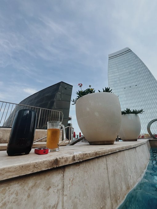 An outdoor rooftop or poolside view showing a black speaker, a glass of beer, and potted succulents on a stone ledge with a modern curved glass skyscraper in the background.