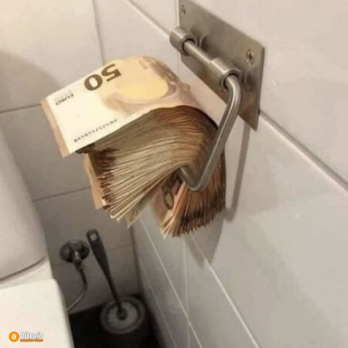 A photograph shows a thick stack of 50 Euro banknotes draped over a metal bathroom toilet paper holder, with a toilet brush visible in the background; a 'bitcoin' logo and watermark are in the bottom left.
