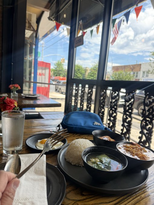 The image shows a dining table with various dishes, including bowls filled with food such as soup or curry. There are also utensils like forks and spoons placed on the table. A person's hand can be seen holding a spoon near one of the bowls. The scene takes place in front of a window that offers a view of a street outside. In the background, there is a car parked along with other cars visible on the street. The image also contains text that reads "This was a really good Thai lunch set. The @useTria card worked at this restaurant. You can get one too and be part of season 3 app.tria.so/?accessCode=VM".