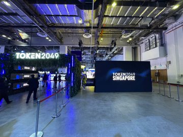 An indoor event space entrance decorated with greenery and blue lighting, featuring large signs reading 'TOKEN2049' and 'TOKEN2049 SINGAPORE'.