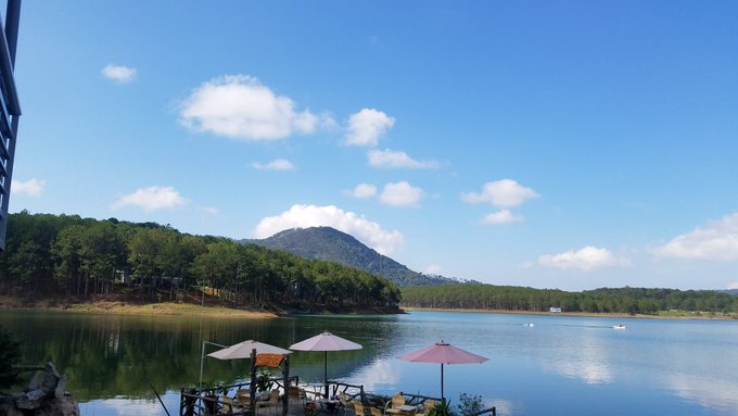 A scenic view of a calm lake surrounded by forested hills and a mountain in the background, with a small dock featuring two umbrellas in the foreground under a blue sky with scattered clouds.