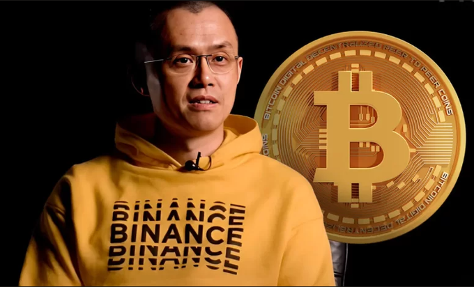 A man wearing glasses and a yellow hoodie printed with 'BINANCE' repeatedly across the chest, standing against a black background with a large golden Bitcoin coin graphic displaying 'BITCOIN DIGITAL... PEER COINS' beside him.