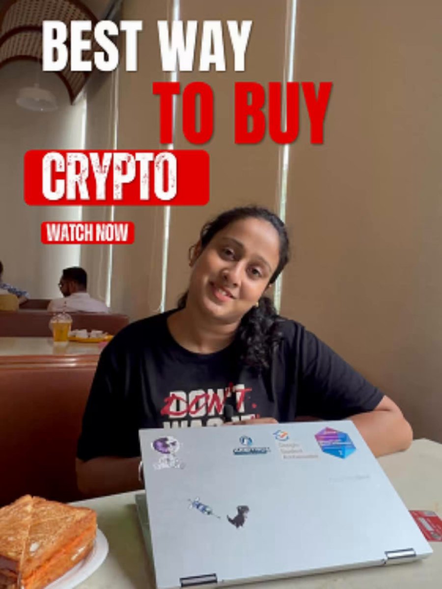 The image features a woman sitting at a table with an open laptop computer, possibly browsing or researching about buying cryptocurrency. The background shows another person seated at a dining table as well. There are also other people present in the scene but they are not the main focus of the image.