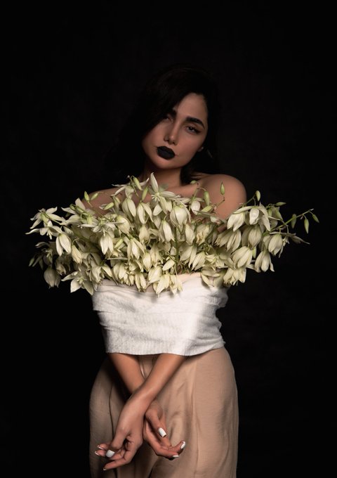 A dark, moody portrait of a woman with dark lipstick holding a bouquet of white flowers across her chest, wearing a white top and beige skirt against a black background.
