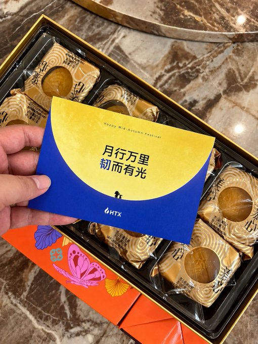 A hand holds a blue and yellow card reading "Happy Mid-Autumn Festival" with Chinese text "月行万里 韧而有光" alongside the HTX (Huobi) logo, set against an elegant gift box of individually wrapped Maxim's mooncakes and a vibrant orange envelope decorated with butterflies, perfectly capturing the festive Mid-Autumn celebration referenced in the caption.
