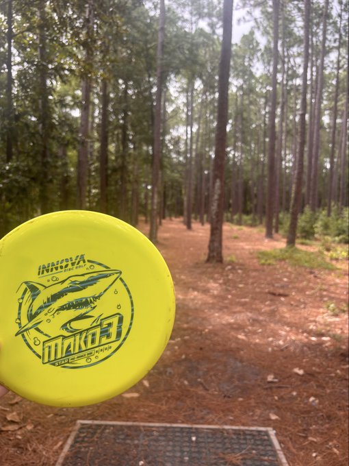 A yellow Innova Star Mako3 disc golf disc held in front of a wooded disc golf course with a metal basket tee pad visible in the background.