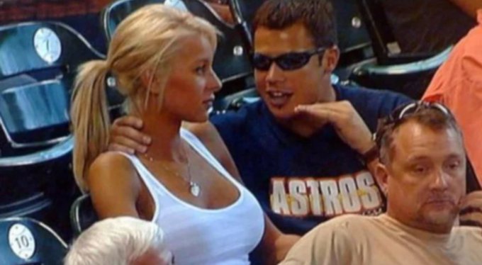 Paris Hilton sits next to a man wearing an Astros jersey and sunglasses in stadium seating, appearing engaged in conversation with him.