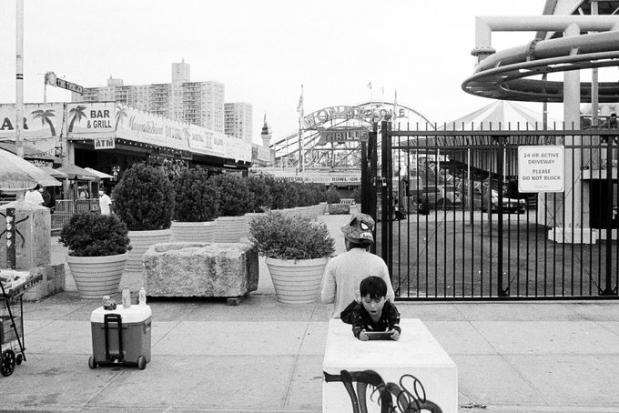 A black-and-white photo shows a child lying on a concrete block and a person in a baseball cap and gray shirt sitting beside him, with an ATM, Bar & Grill signs, a roller coaster, and an "Astroland" sign visible in the background, with the "24 HR ACTIVE DRIVEWAY PLEASE DO NOT BLOCK" gate sign on the right. The image's NYC seaside amusement park setting, captured in candid street photography style, reflects the storytelling approach behind the auctioned piece from the Archive: NYC (2022) collection, where everyday urban moments become meaningful works of art.