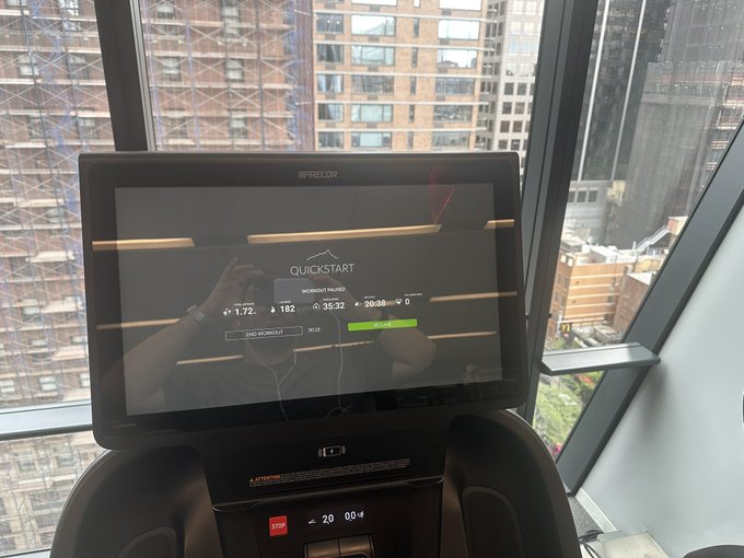 View of a Precor treadmill console in 'Quickstart' mode in a high-rise gym, with large windows showing a cityscape of tall buildings in the background.