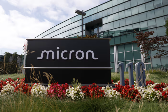 A black sign reading "micron" in white lowercase letters stands in front of a modern glass building, surrounded by a flower bed with red and white blooms and a "10" sculpture.
