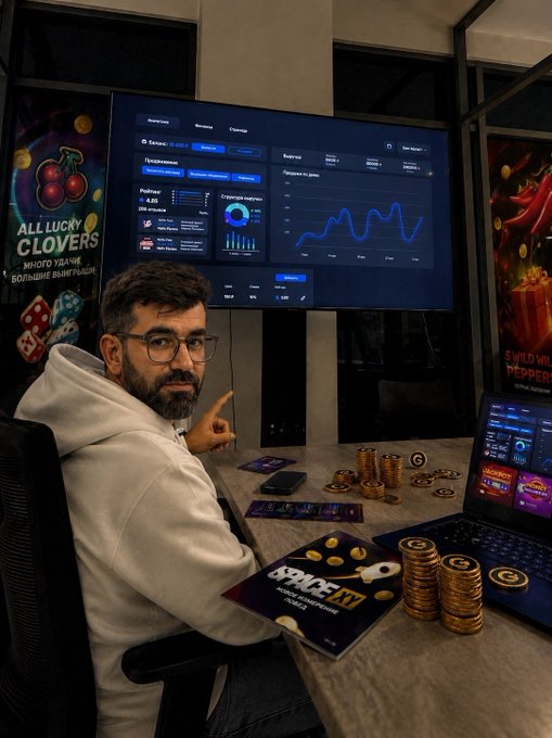 A bearded man with glasses in a white hoodie sits at a desk pointing toward a large monitor displaying gaming analytics dashboards, with stacks of gold coins and "SPACE XVII" promotional materials nearby, alongside a laptop showing the Playnance ecosystem interface. The setup visually represents the expanding Playnance Web3 gaming platform highlighted in the post about G Coin's rapid growth.
