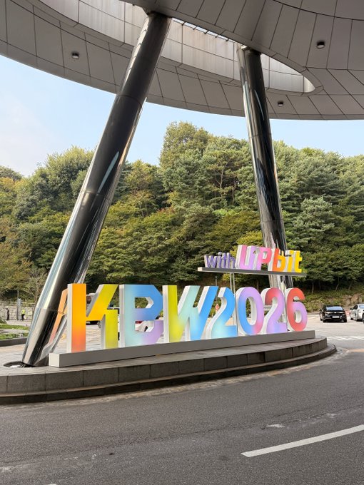 A colorful 'KBW 2026 with UPbit' sign displayed outdoors under a modern architectural canopy with trees in the background.