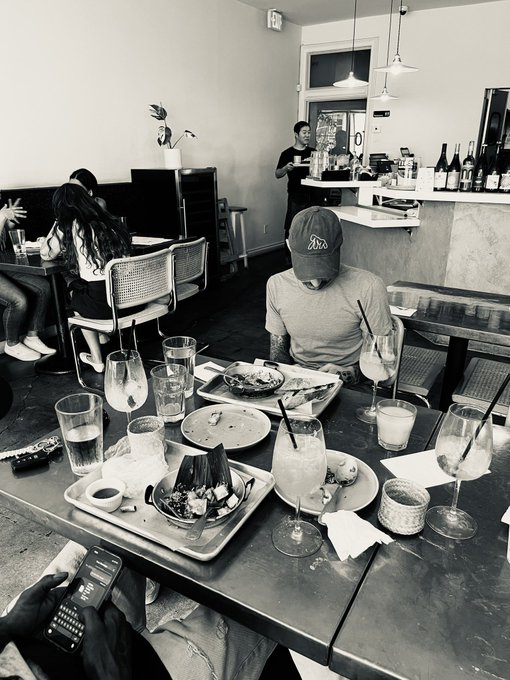 A man in a cap sits at a cluttered restaurant table full of empty plates, water glasses, and cocktails, while friends dine in the background. The scene captures the post-meal aftermath of a casual brunch outing between close friends.