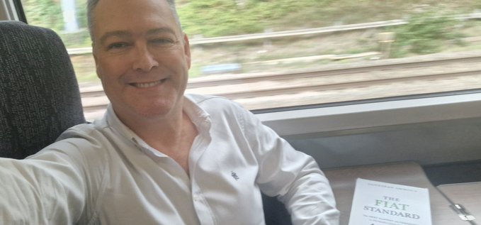 A smiling man in a white button-down shirt takes a selfie while sitting on a train, with a book titled 'THE FAT STANDARD' visible on the seat beside him.